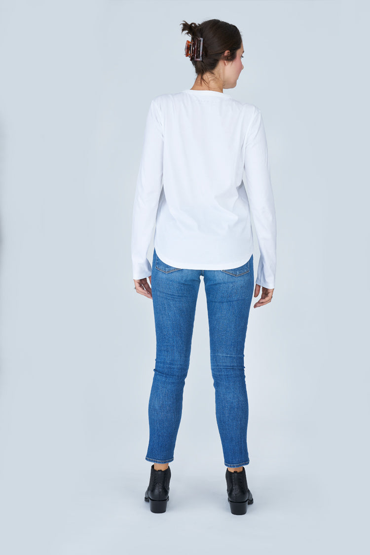 A woman faces away in a plain light gray setting, wearing The Catherine 100% cotton crew neck tee by SUZI ROHER with blue jeans and black ankle boots. Her hair is tied up with a large brown clip.