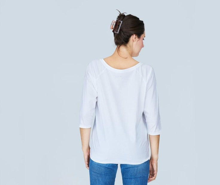 A woman with dark hair in a clip, wearing SUZI ROHER’s The Bailey boxy cut white tee with three-quarter sleeves and blue jeans, stands facing away from the camera against a light blue background.