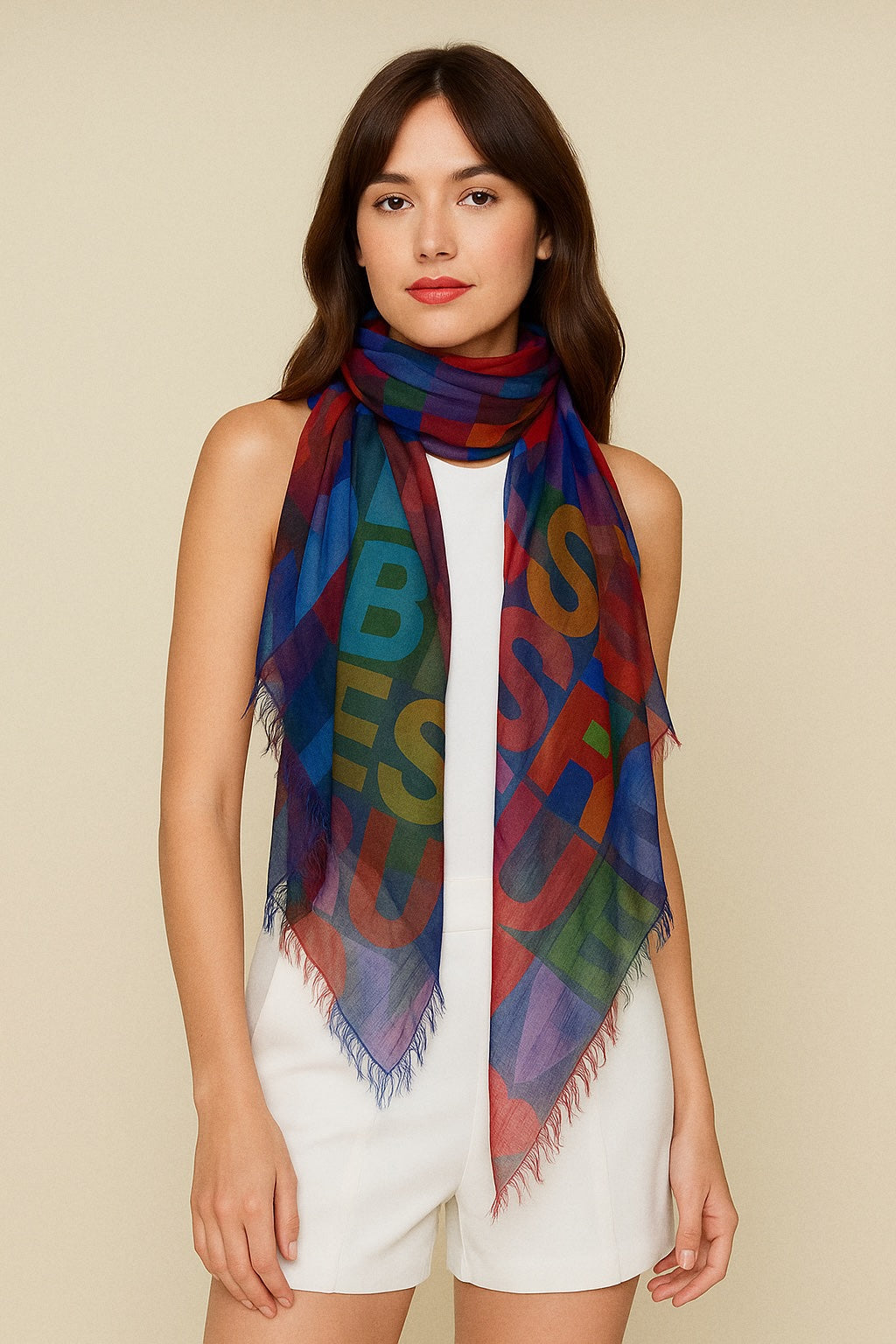 A woman with long brown hair wears a sleeveless white romper and the TRUE affirmation scarf by SUZI ROHER, featuring large multicolored letters, as she stands against a plain beige background.