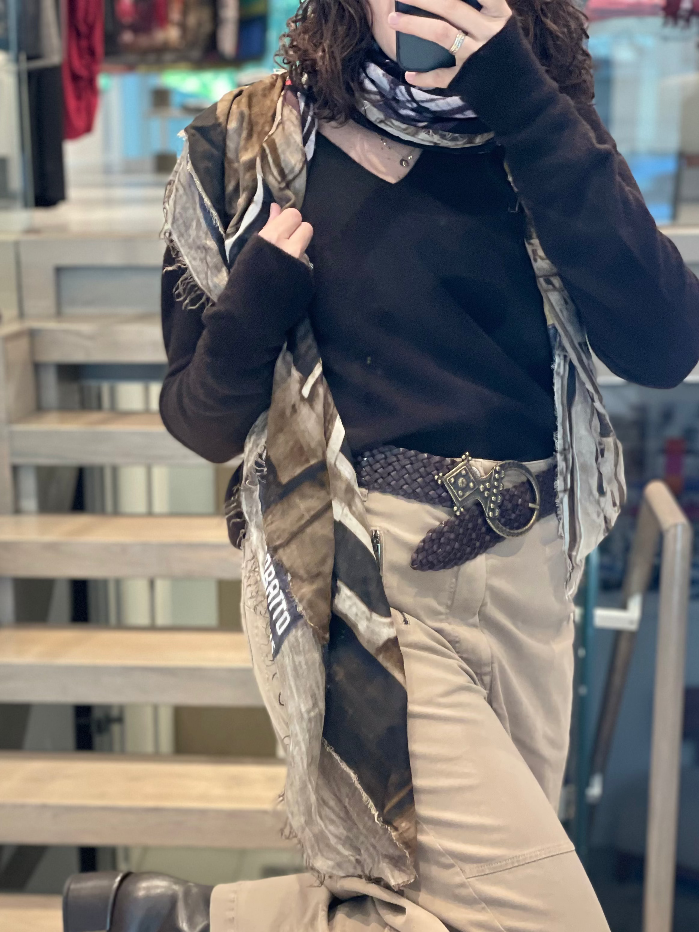A person stands on stairs wearing the SUZI ROHER 8 1/2 black long-sleeve top, beige pants, a wide woven belt, and a cinematic scarf partially covering their face as they take a mirror selfie.