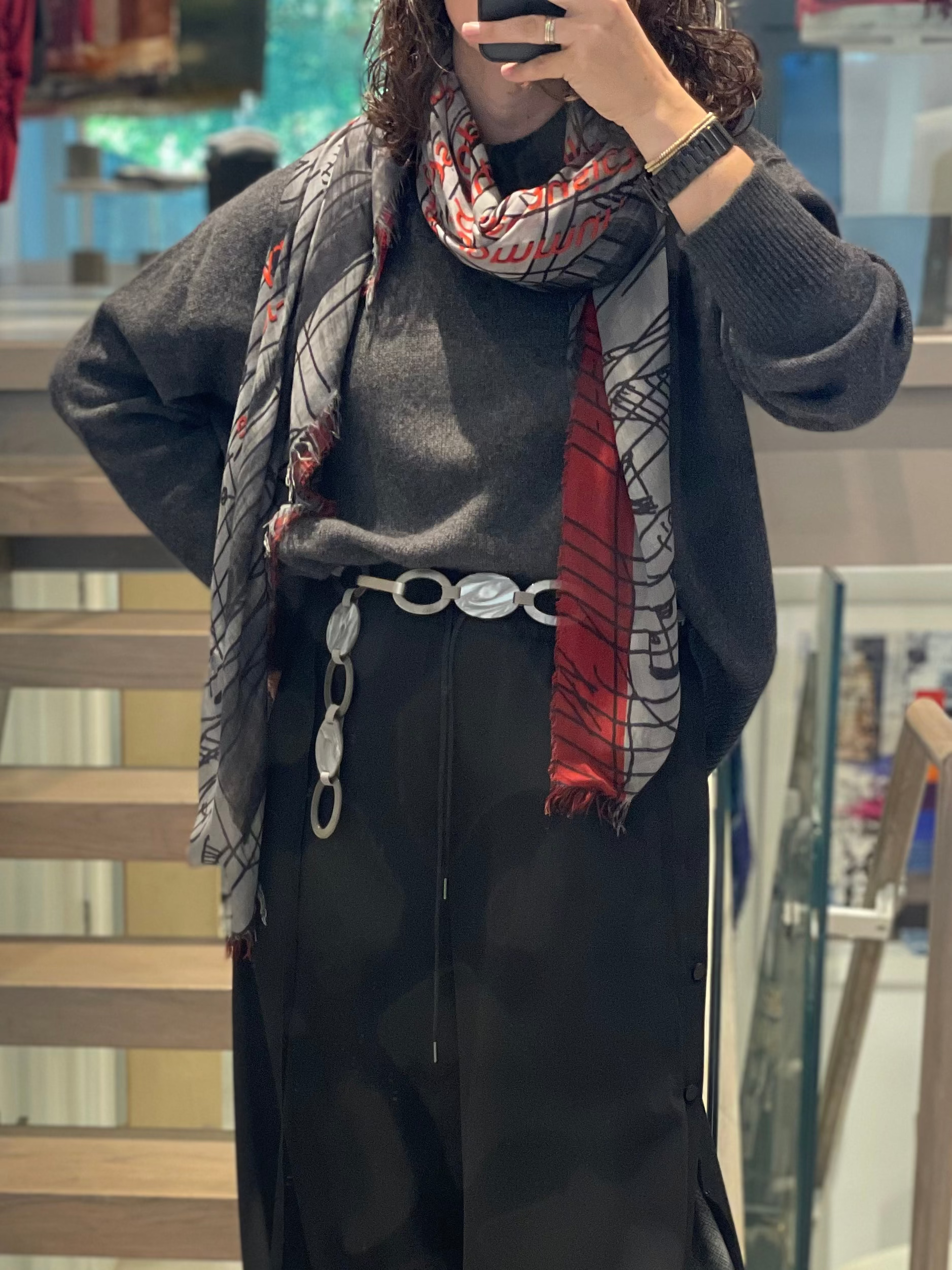Showcasing artistic fashion, a person in a gray sweater, black skirt with wide silver chain belt, smartwatch, and a SUZI ROHER John Cage scarf with red accents poses for an indoor mirror selfie.
