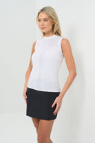 Side view of a sleeveless white mock-neck knit top featuring delicate pointelle detailing across the upper chest and a vertical textured stripe down the center front. Paired with black shorts for a sleek, polished look.