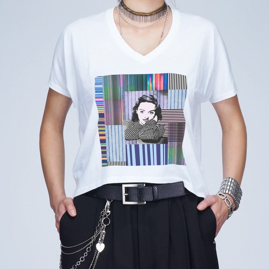 A person wears The Velvet tee by SUZI ROHER, featuring a Bette Davis graphic over a colorful abstract background, styled with layered necklaces, bracelets, a belt, and black pants.