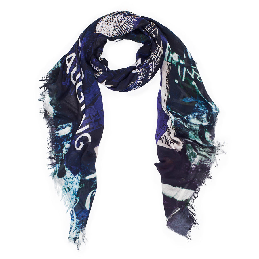 The SUZI ROHER Madame Fortuna Vol 2 scarf is a dark, rectangular modal silk piece with frayed edges, abstract patterns, text like "Love Fate and Destiny," and hues of navy blue, green, white, and black, styled in a loose loop.