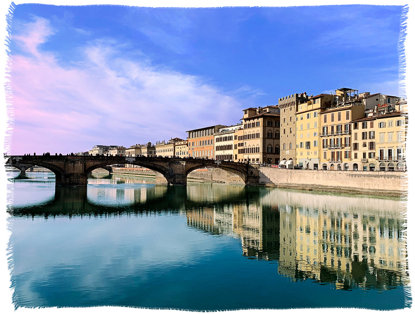 A stone bridge over a calm river reflects vibrant historic buildings and a blue sky—evoking the charm of enjoying SUZI ROHER’s Kim Campari Time aperitif along Italy’s scenic waterways.