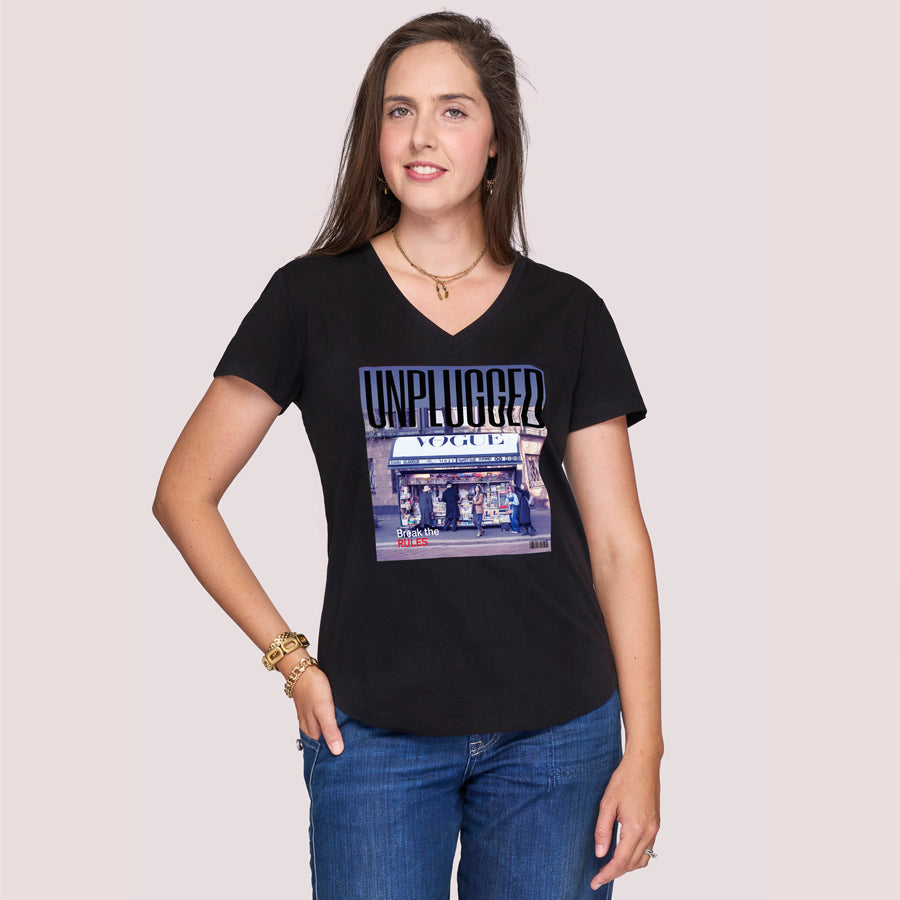 A woman with long brown hair, in blue jeans and gold jewelry, smiles gently against a light background, wearing SUZI ROHER’s “The Viv” slim fit black V-neck tee featuring a Vogue-style image on the front.