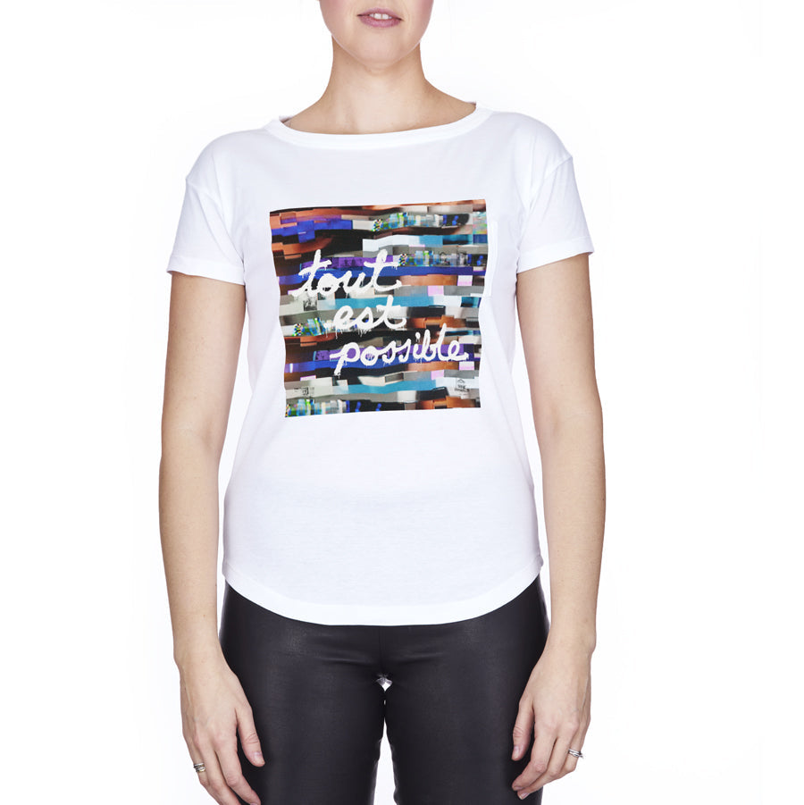A woman wears the SUZI ROHER "Brie Tout Est Possible" white t-shirt with a vibrant graphic and inspiring script, paired with black pants, standing against a plain white background, embodying the spirit of possibility.