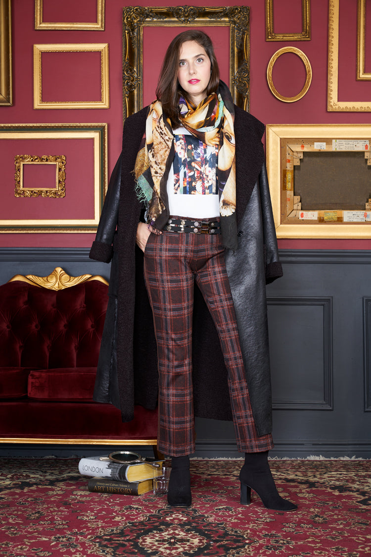 A woman wearing The Catherine by SUZI ROHER, a long black coat paired with plaid pants and heeled boots, stands confidently before an ornate red and gold Renaissance-style wall, next to a red velvet sofa and stacked books on the floor.