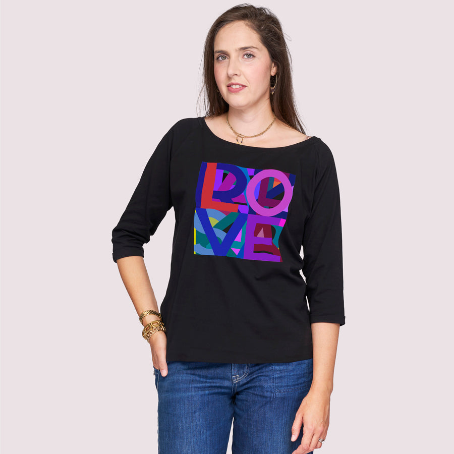 A woman with long brown hair wears SUZI ROHER’s The Bailey, a black boxy cut tee featuring a colorful geometric LOVE graphic, paired with blue jeans and standing against a plain light background.
