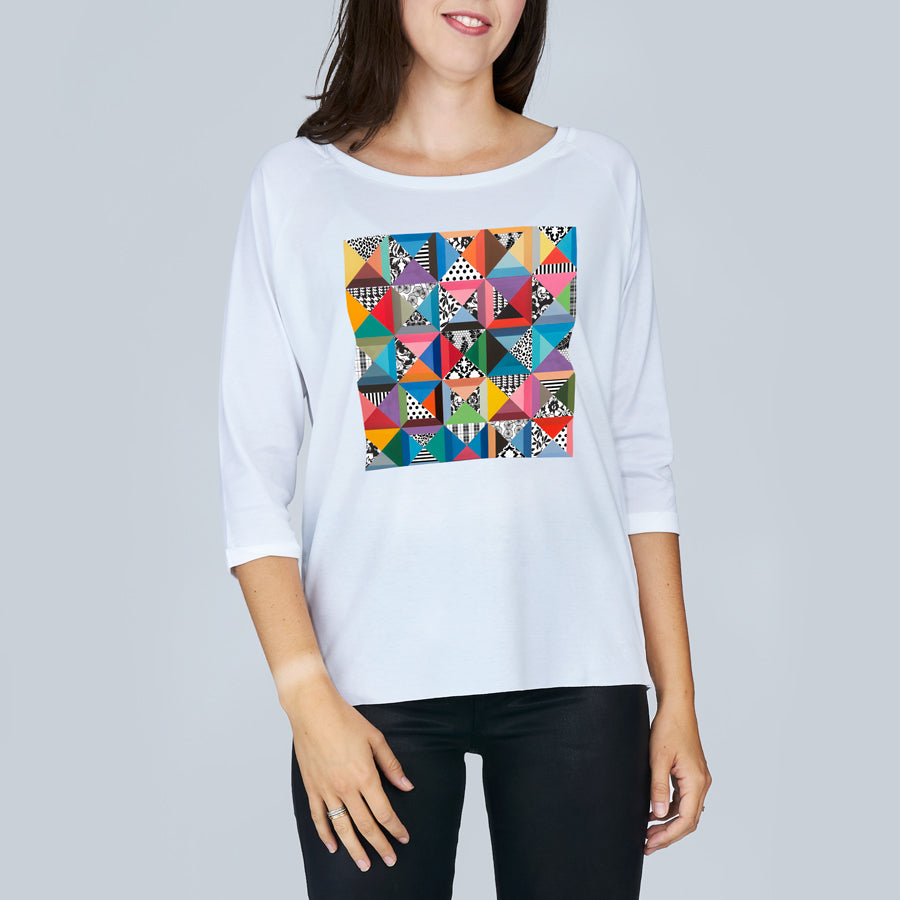 A woman models The Bailey by SUZI ROHER—a white, oversized boxy tee featuring a vibrant geometric patchwork of triangles and unique designs—standing against a plain light gray background.