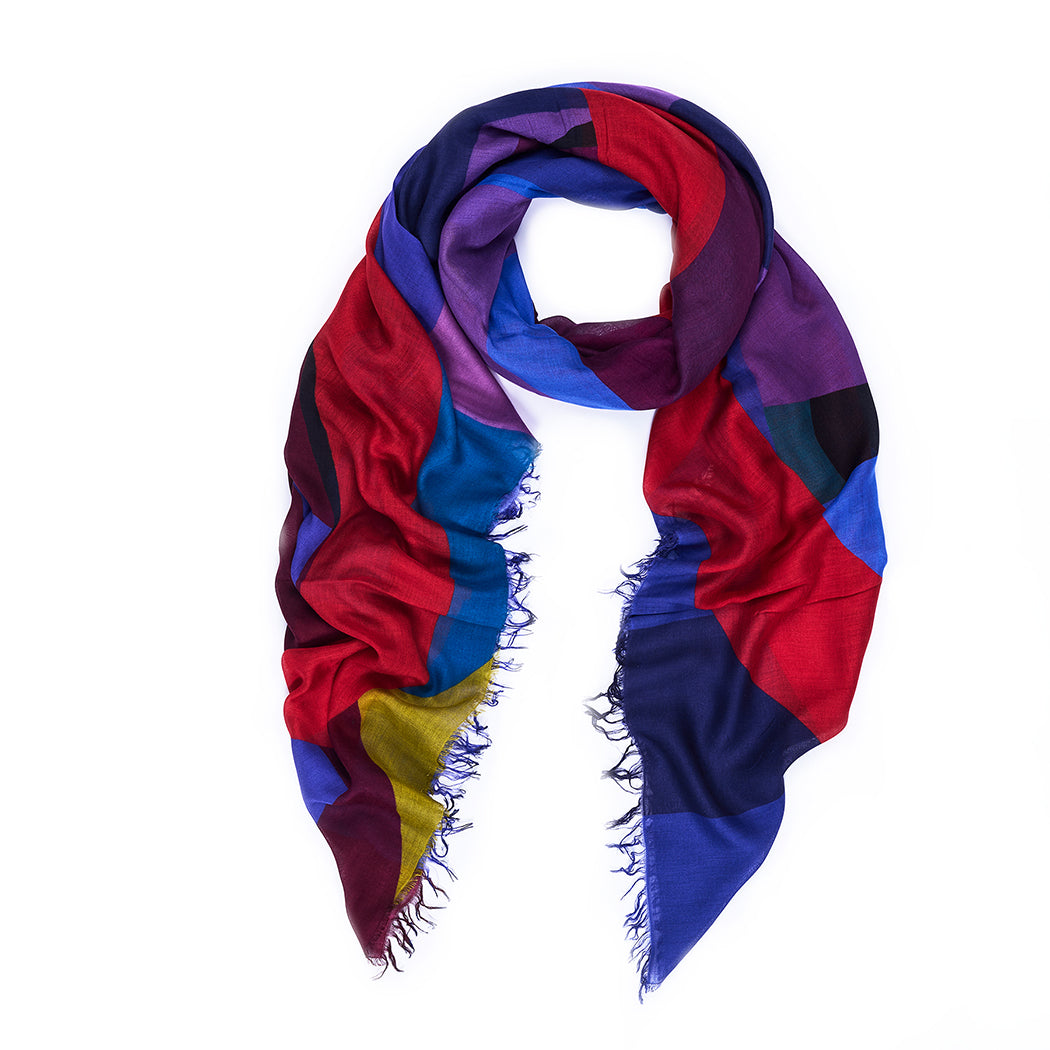 The SUZI ROHER CIAO scarf features vibrant abstract geometric patterns in red, blue, purple, yellow, and black, with frayed edges and bold letters inspired by an Italian song. It’s displayed on a white background.