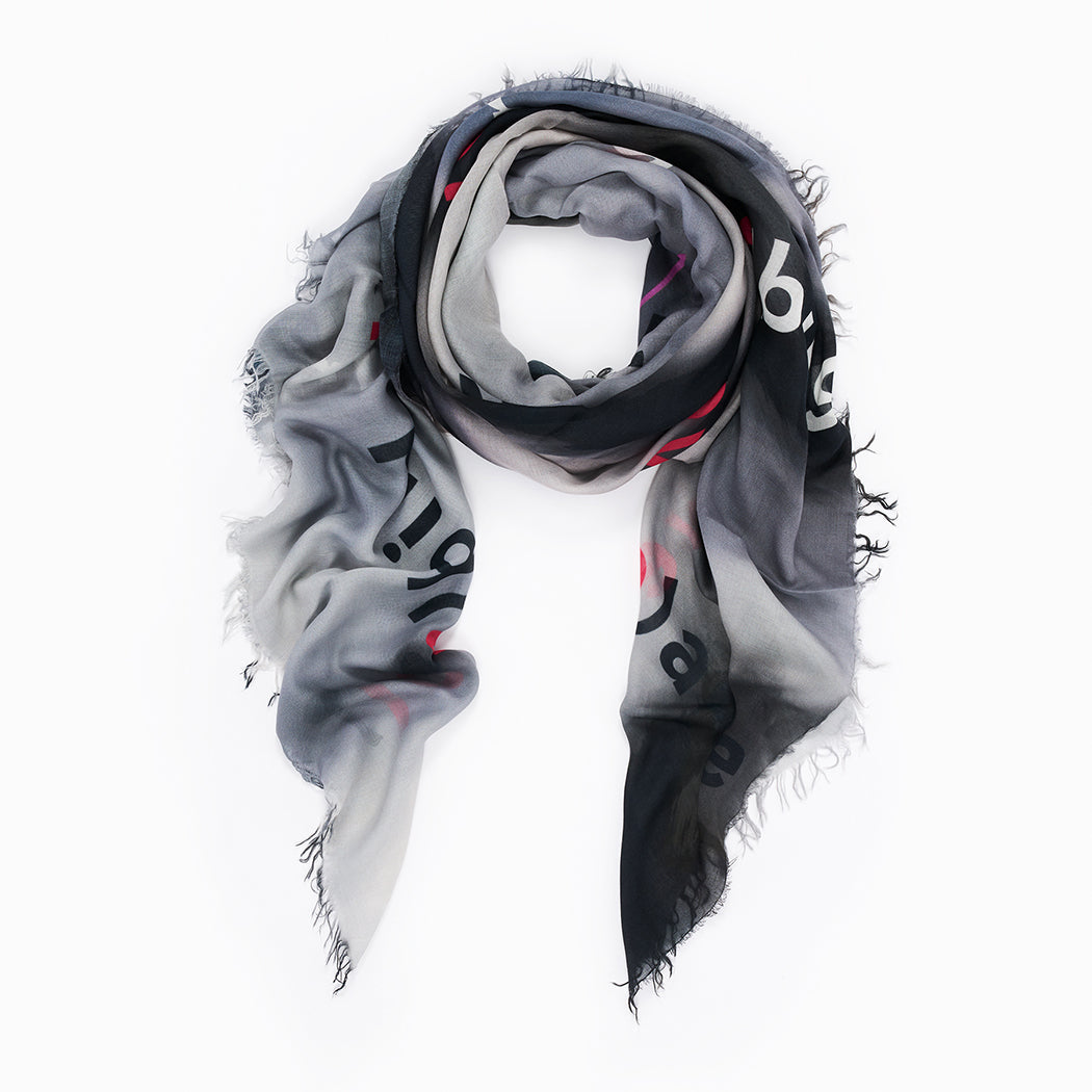 The SUZI ROHER "Lovefest" scarf is a lightweight modal silk accessory with frayed edges, featuring a black, gray, and white gradient and bold black and red letters spelling "love." Shown loosely coiled on a white background.