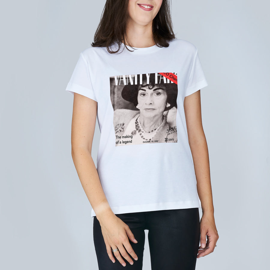 A woman looks fabulous in SUZI ROHER's The Niki, a classy white T-shirt with a black-and-white photo like a magazine cover and the words VANITY and The making of a legend, smiling against a plain background.