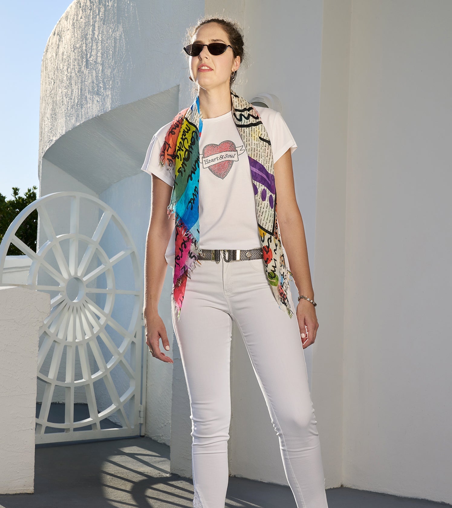 A woman wearing SUZI ROHER's The Karlie—a bedazzled white graphic tee—pairs it with white pants, sunglasses, and a colorful scarf as she stands confidently before a modern white building with geometric lines.