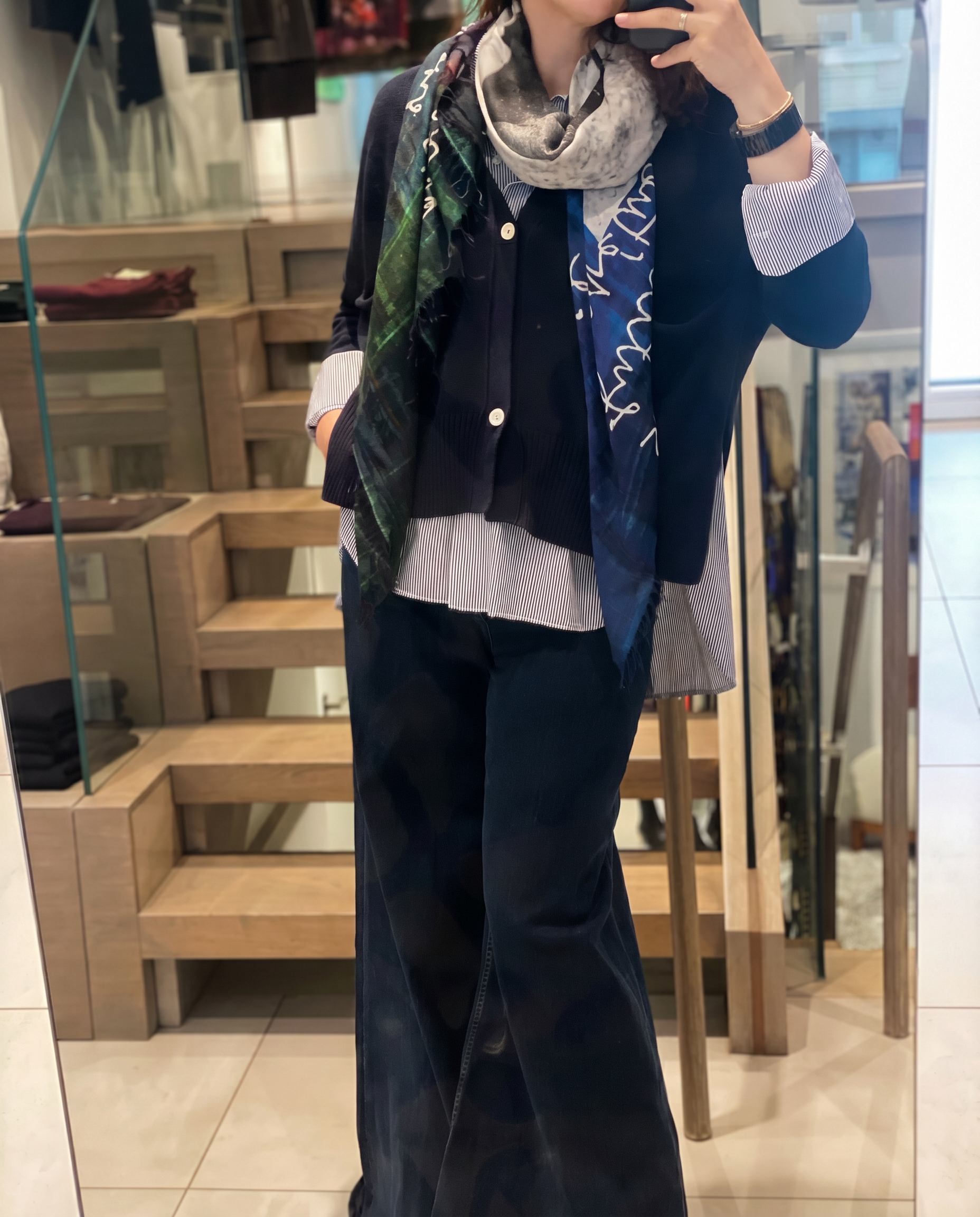 A person snaps a mirror selfie in a store, styled in SUZI ROHER’s A La Mode silk scarf—featuring green, blue, and gray patterns—paired with wide-leg dark pants, a dark blazer, and striped shirt. Their face is hidden by the phone.