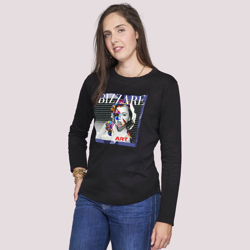 A woman with long brown hair wears SUZI ROHER’s The Catherine, a black 100% cotton crew-neck long-sleeve shirt featuring a colorful “BIZZARE” graphic with a face, styled with blue jeans against a light background.