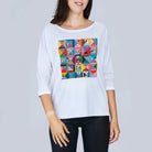 A woman wears The Bailey by SUZI ROHER, a white boxy tee with 3/4 sleeves and a vibrant geometric quilt pattern on the front, standing against a plain light gray background.