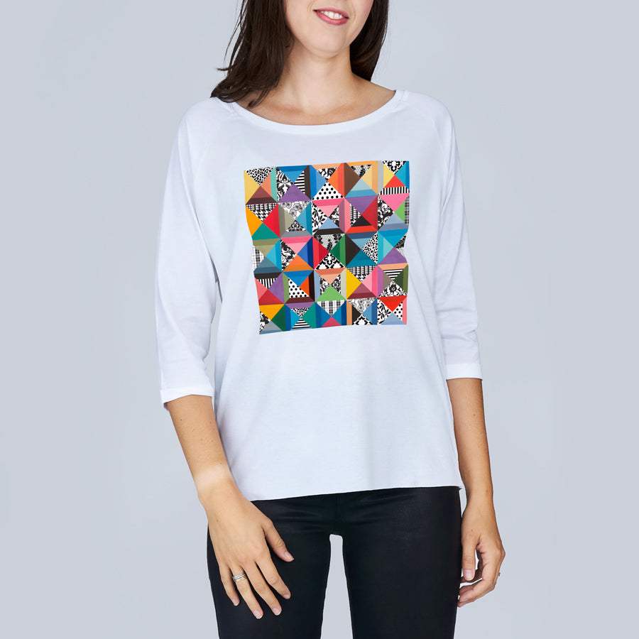A woman wears The Bailey by SUZI ROHER, a white boxy tee with 3/4 sleeves and a vibrant geometric quilt pattern on the front, standing against a plain light gray background.