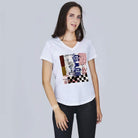 A woman with long brown hair wears SUZI ROHER's The Viv, a white V-neck tee with a colorful abstract print, paired with black pants, standing against a plain light gray background.