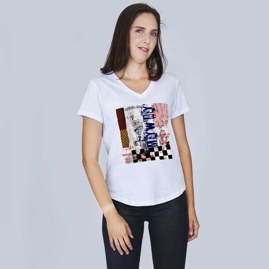 A woman with long brown hair wears SUZI ROHER's The Viv, a white V-neck tee with a colorful abstract print, paired with black pants, standing against a plain light gray background.