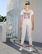 A woman wearing The Viv by SUZI ROHER—sunglasses, a white graphic tee, white pants, and sneakers—stands stylishly outdoors beside a white building and large wheel as sunlight casts striking shadows.