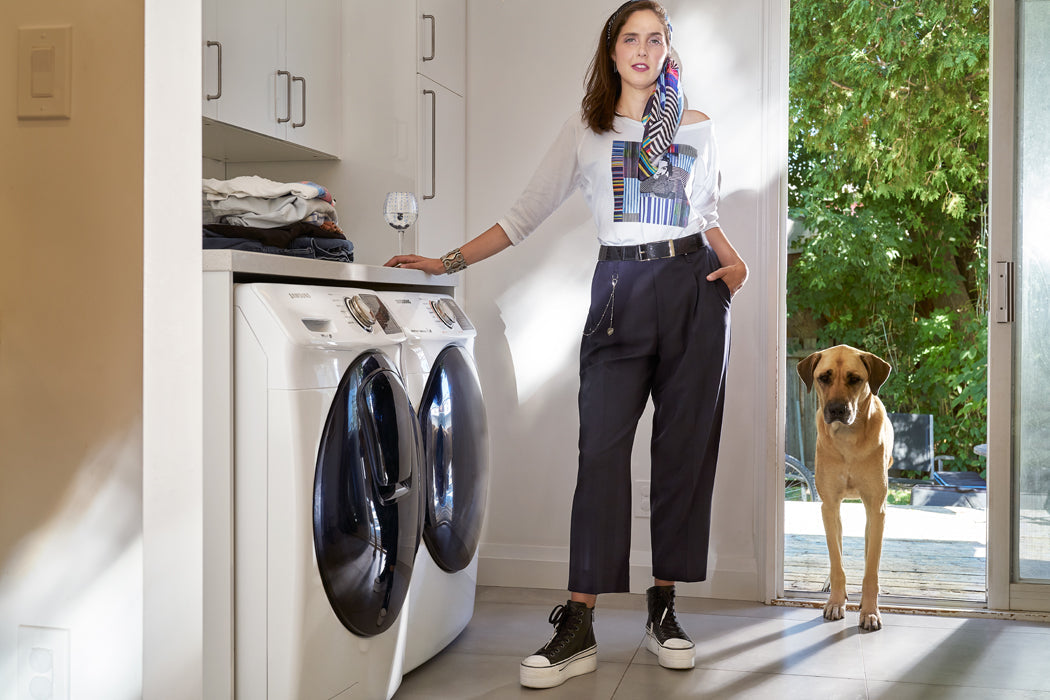 Wearing SUZI ROHER's The Bailey boxy cut tee, a woman stands in a laundry room with dark pants and sneakers, holding wine beside a washer and dryer as her large dog waits in the doorway to a sunlit green yard.