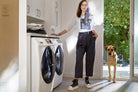 Wearing SUZI ROHER's The Bailey boxy cut tee, a woman stands in a laundry room with dark pants and sneakers, holding wine beside a washer and dryer as her large dog waits in the doorway to a sunlit green yard.