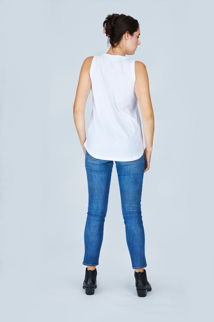 A woman faces away against a light grey background, wearing SUZI ROHER’s The Lola sleeveless white 100% cotton tank top, paired with blue jeans and black ankle boots.