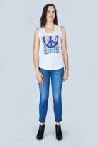 A woman stands against a light gray background, wearing SUZI ROHER’s The Lola— a 100% cotton white tank top with a blue peace symbol—paired with blue jeans and black ankle boots, looking forward with arms relaxed at her sides.