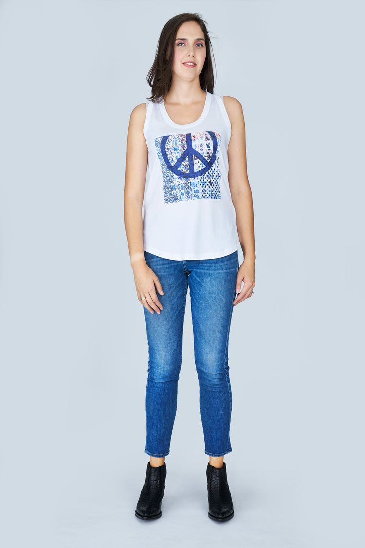 A woman stands against a light gray background, wearing SUZI ROHER’s The Lola— a 100% cotton white tank top with a blue peace symbol—paired with blue jeans and black ankle boots, looking forward with arms relaxed at her sides.