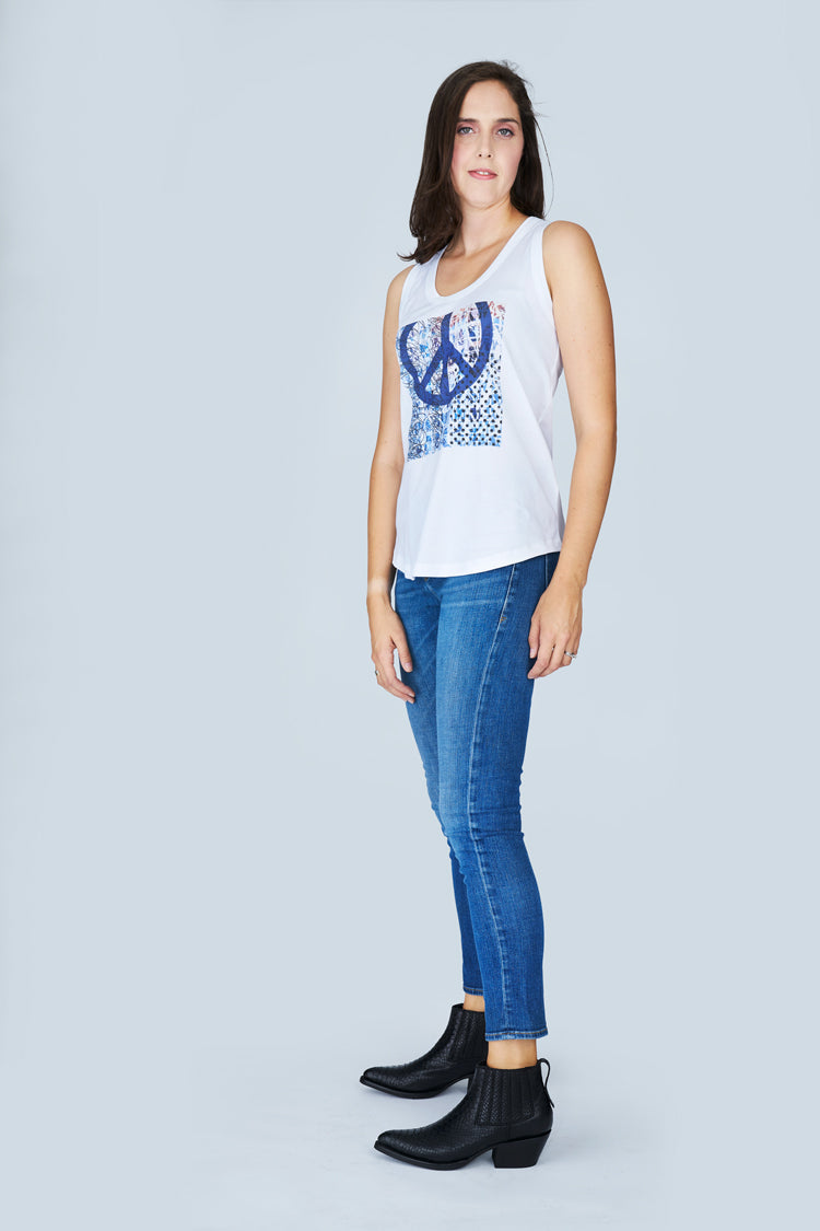 A woman with straight brown hair models The Lola white sleeveless tank top by SUZI ROHER, featuring a blue peace symbol. She pairs it with blue jeans and black ankle boots, posing against a plain light gray background.