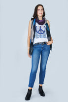A woman stands against a light background, wearing The Lola by SUZI ROHER: a 100% cotton white tank top with a blue peace symbol, paired with blue jeans, a multicolored scarf, black ankle boots, and a black belt. She has one hand in her pocket and smiles slightly.