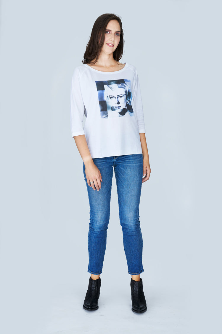 A woman with long brown hair wears The Bailey by SUZI ROHER—a white boxy-cut tee with 3/4 sleeves and a graphic print—paired with blue jeans and black ankle boots, standing against a light grey background.