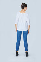 A woman stands facing away, wearing SUZI ROHER's The Bailey white boxy cut tee, blue skinny jeans, and black ankle boots. Her hair is clipped up against a light gray background for timeless Audrey Hepburn-inspired elegance.