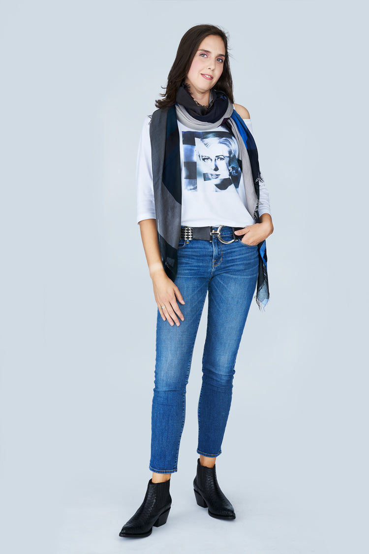 A woman with shoulder-length dark hair wears SUZI ROHER's The Bailey, a boxy-cut 3/4 sleeve tee, styled with blue jeans, black ankle boots, a black belt, and a black-and-white portrait print scarf. She stands facing forward with one hand in her pocket.