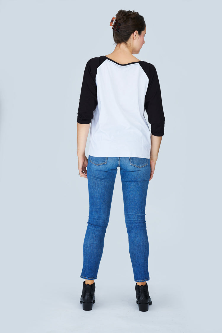 A woman with her hair tied up stands facing away against a light gray background, wearing SUZI ROHER’s The Bailey tee—100% cotton, white with black boxy cut and “There is Hope” graphic—paired with blue jeans and black ankle boots.