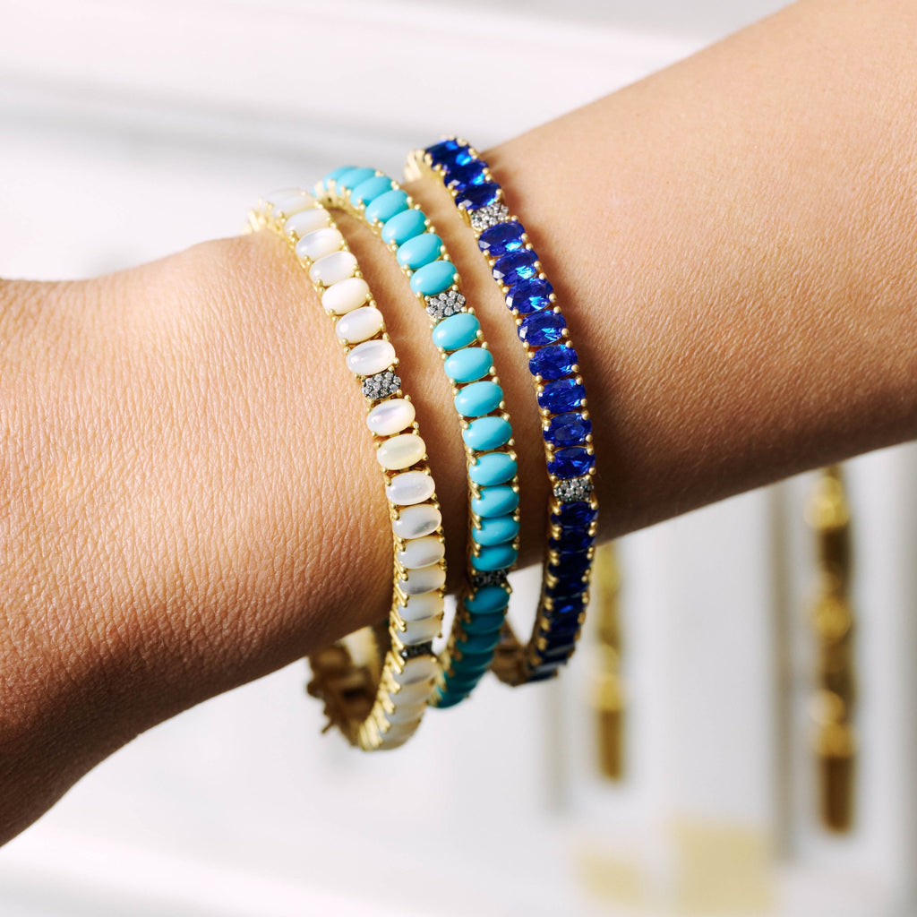 A close-up image of a person's wrist adorned with three Midnight Tri-Tone Twilight Oval Station Tennis Bracelets by Midnight. The bracelets feature 14K matte gold settings and are embedded with oval-shaped gems in white, turquoise, and deep blue colors, each interspersed with sparkling cubic zirconia stones.