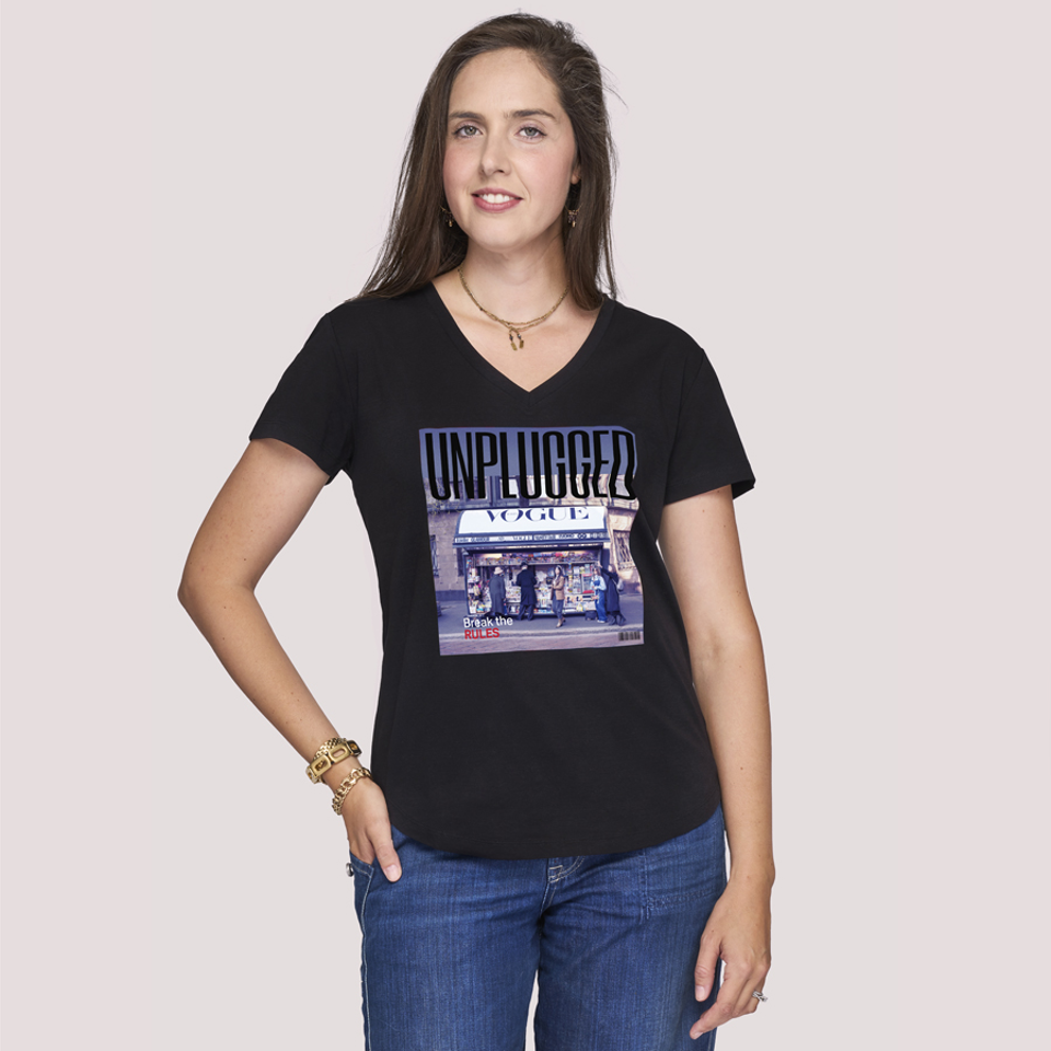 A woman with long brown hair smiles against a light background, one hand in her pocket, wearing SUZI ROHER's The Viv—a black 100% cotton V-neck tee with a vibrant UNPLUGGED VOGUE graphic on the front.