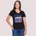 A woman with long brown hair smiles slightly as she poses against a light plain background, wearing The Viv slim-fit black V-neck tee by SUZI ROHER with a Vogue UNPLUGGED graphic, paired with blue jeans and gold jewelry.
