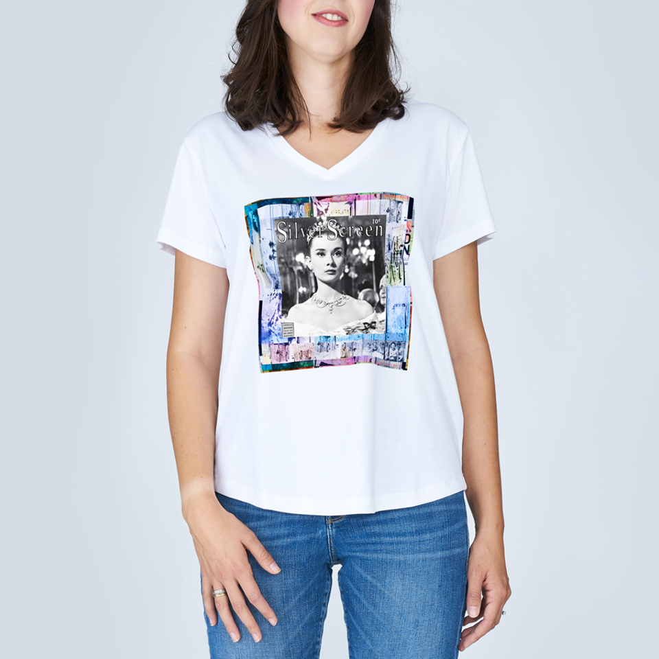 A woman wears The Viv by SUZI ROHER—a white V-neck T-shirt with a black-and-white Audrey Hepburn photo framed by a colorful border—paired with blue jeans, standing against a light gray background.