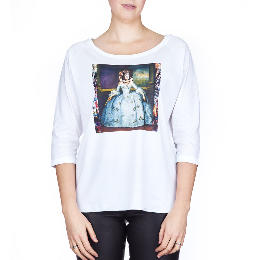 A woman stands against a white background wearing SUZI ROHER’s The Bailey—a white, boxy-cut tee with a boat neckline, featuring a historical painting print of a woman in a blue dress on the front.