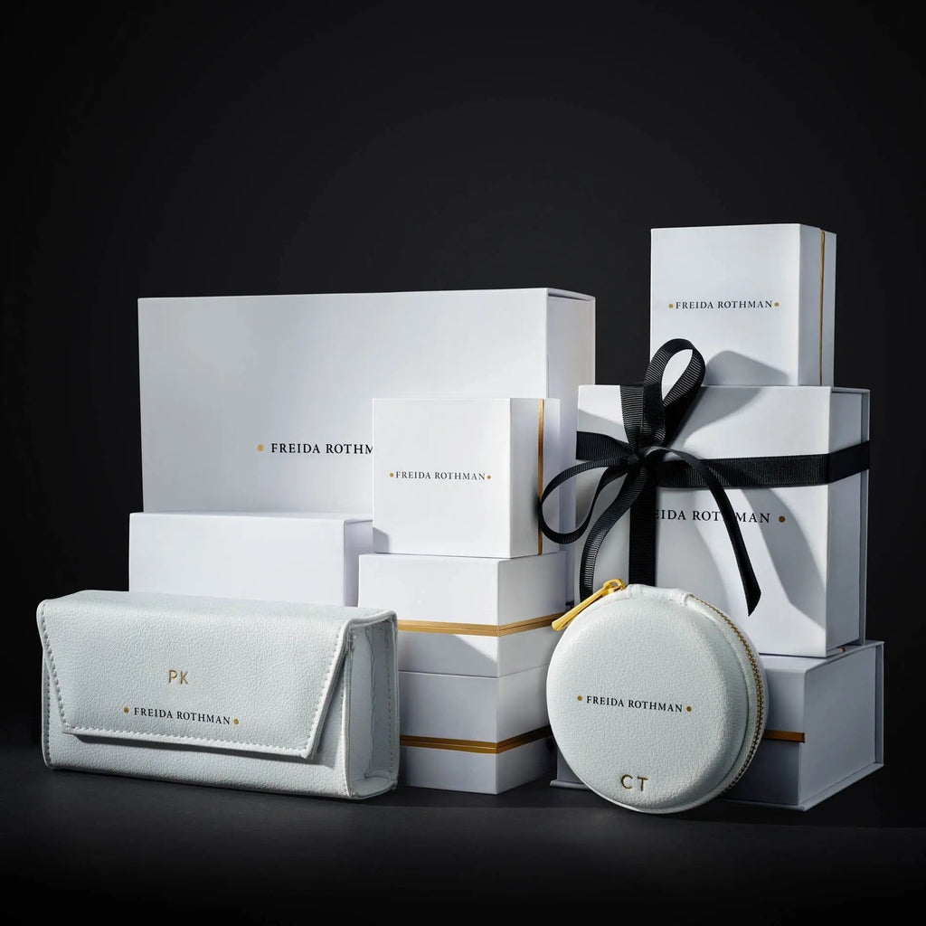 A collection of white boxes with gold accents and the brand name "FREIDA ROTHMAN" displayed on them. In front are a light gray wallet-type pouch labeled "PK" and a round zip pouch labeled "CT." Some boxes, adorned with black ribbons, hint at treasures like the Pavé Thin Marquee Ring in sterling silver or 14K gold inside.