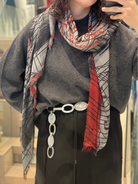 A person wears a loose gray sweater, a black skirt, and a belt with large white chain links, styled with the SUZI ROHER "John Cage" scarf featuring minimalist gray and red patterns. Their hand rests on their hip; part of their face is out of frame.