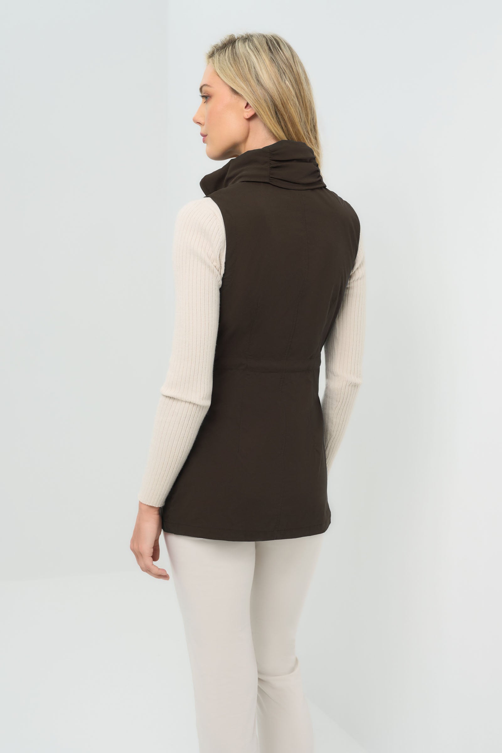 Back view of an espresso-colored vested jacket, zipped up with a collar, layered over a white zip-up hoodie and paired with white slim-fit pants.
