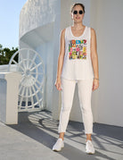 A woman stands outside in sunlight near a white building featuring circular decor, wearing The Lola graphic tank top by SUZI ROHER, paired with sunglasses, white pants, and white sneakers.
