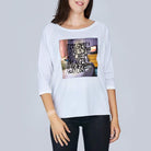 A woman is smiling while wearing SUZI ROHER’s “The Bailey” white long-sleeve shirt with a graphic and the text: "It was great. I love the coffee with charm and such. Coffee In Bed was great! Not love." She stands against a plain background.