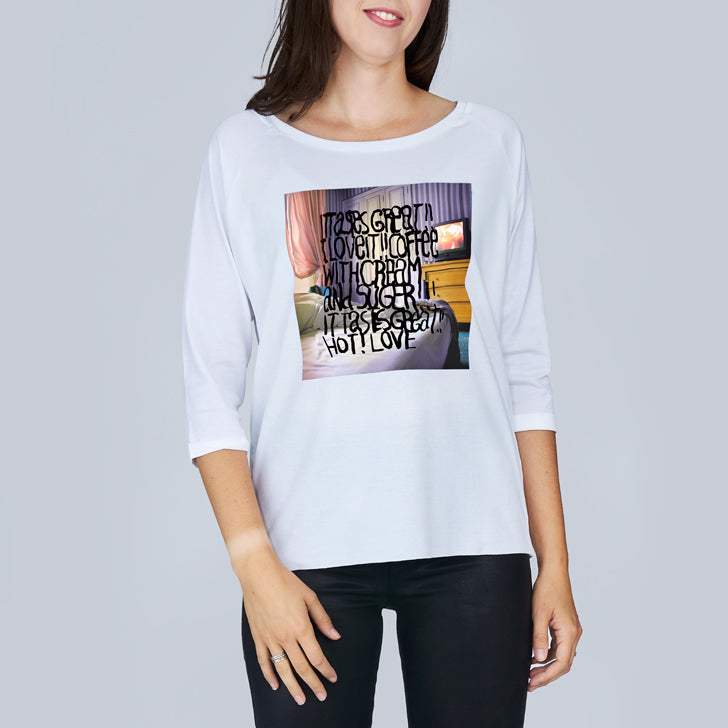 A woman is smiling while wearing SUZI ROHER’s “The Bailey” white long-sleeve shirt with a graphic and the text: "It was great. I love the coffee with charm and such. Coffee In Bed was great! Not love." She stands against a plain background.