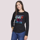 A woman with long brown hair wears The Catherine crew neck tee by SUZI ROHER, featuring a colorful graphic, paired with blue jeans. She stands against a plain light background, looking at the camera with a slight smile.