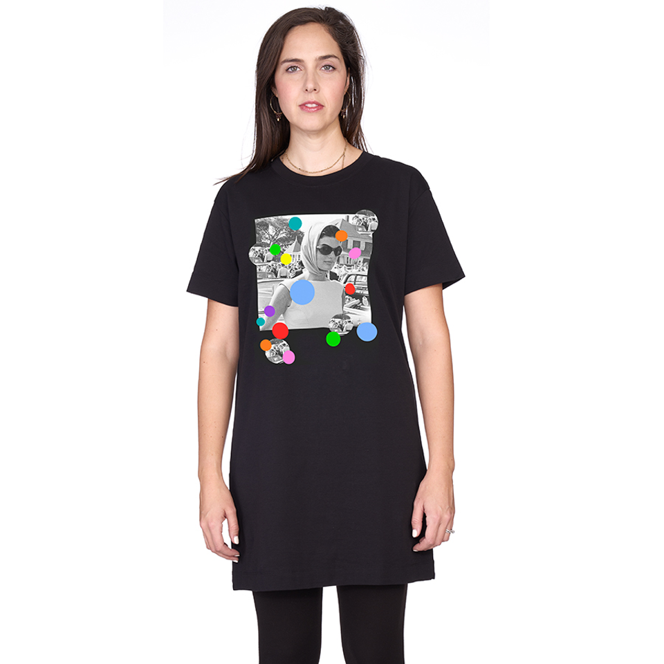 A woman with long brown hair wears the oversized "Daisy, Because We love You" black t-shirt dress by SUZI ROHER, featuring a grayscale collage graphic with colorful circles on the front. She stands against a plain white background.
