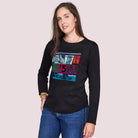 A woman with long brown hair models The Catherine by SUZI ROHER—a black 100% cotton crew neck long-sleeve tee with a colorful $5 captcha graphic—paired with blue jeans, against a plain light background, looking at the camera.